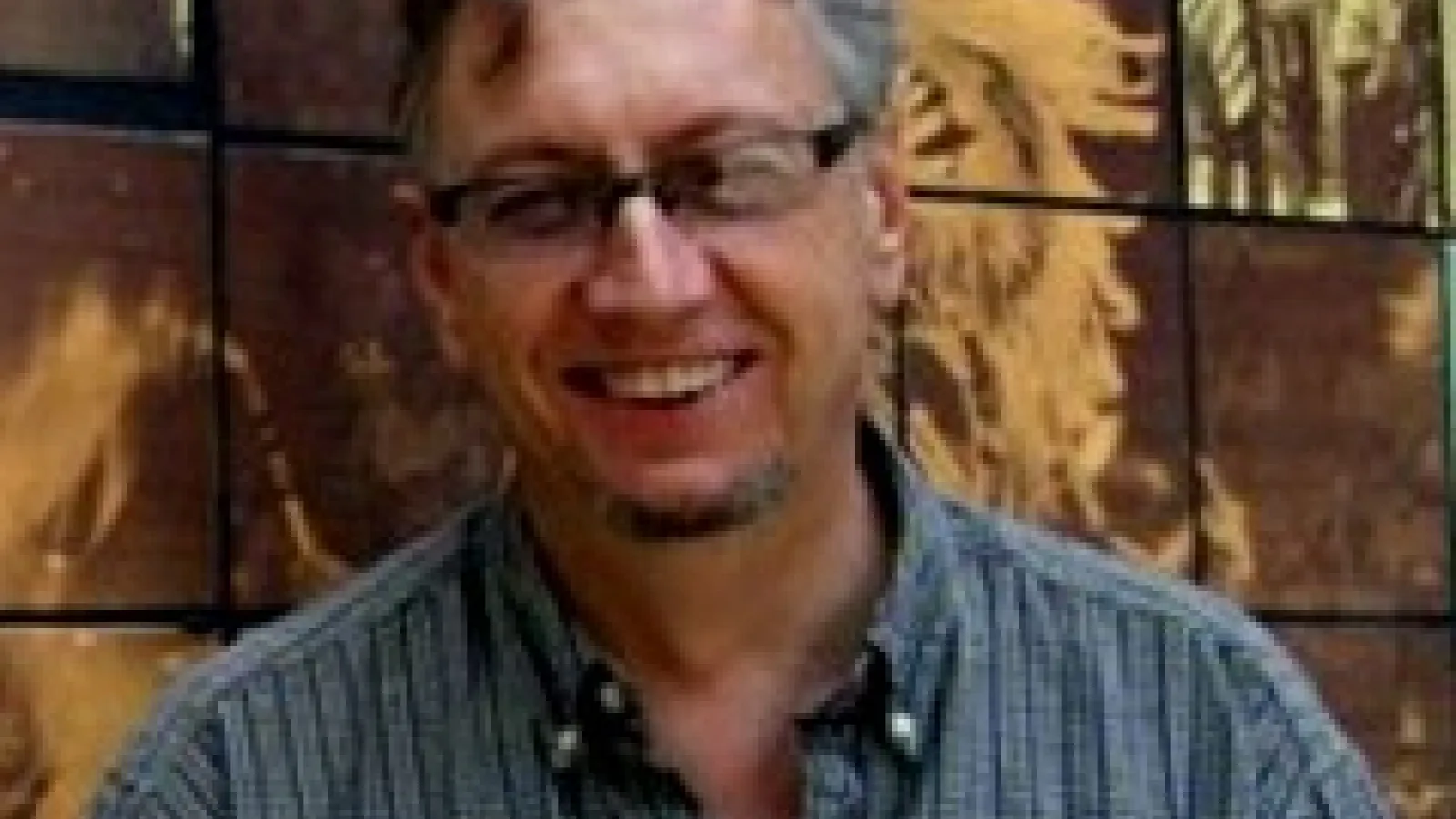 John Low in a blue shirt and glasses in front of a mosaic wall.