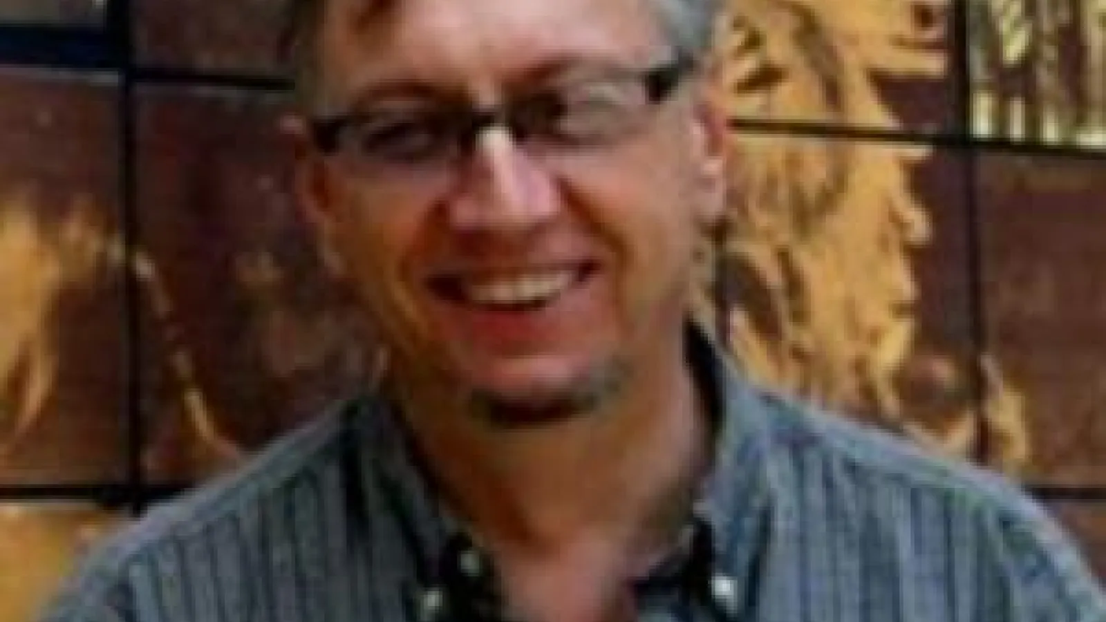John Low in a blue shirt and glasses in front of a mosaic wall.