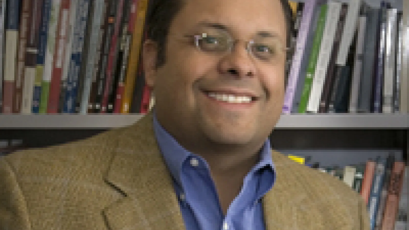 Bryan Brayboy, a tan-skinned man with glasses and a brown jacket sitting in front of a bookcase, smiling