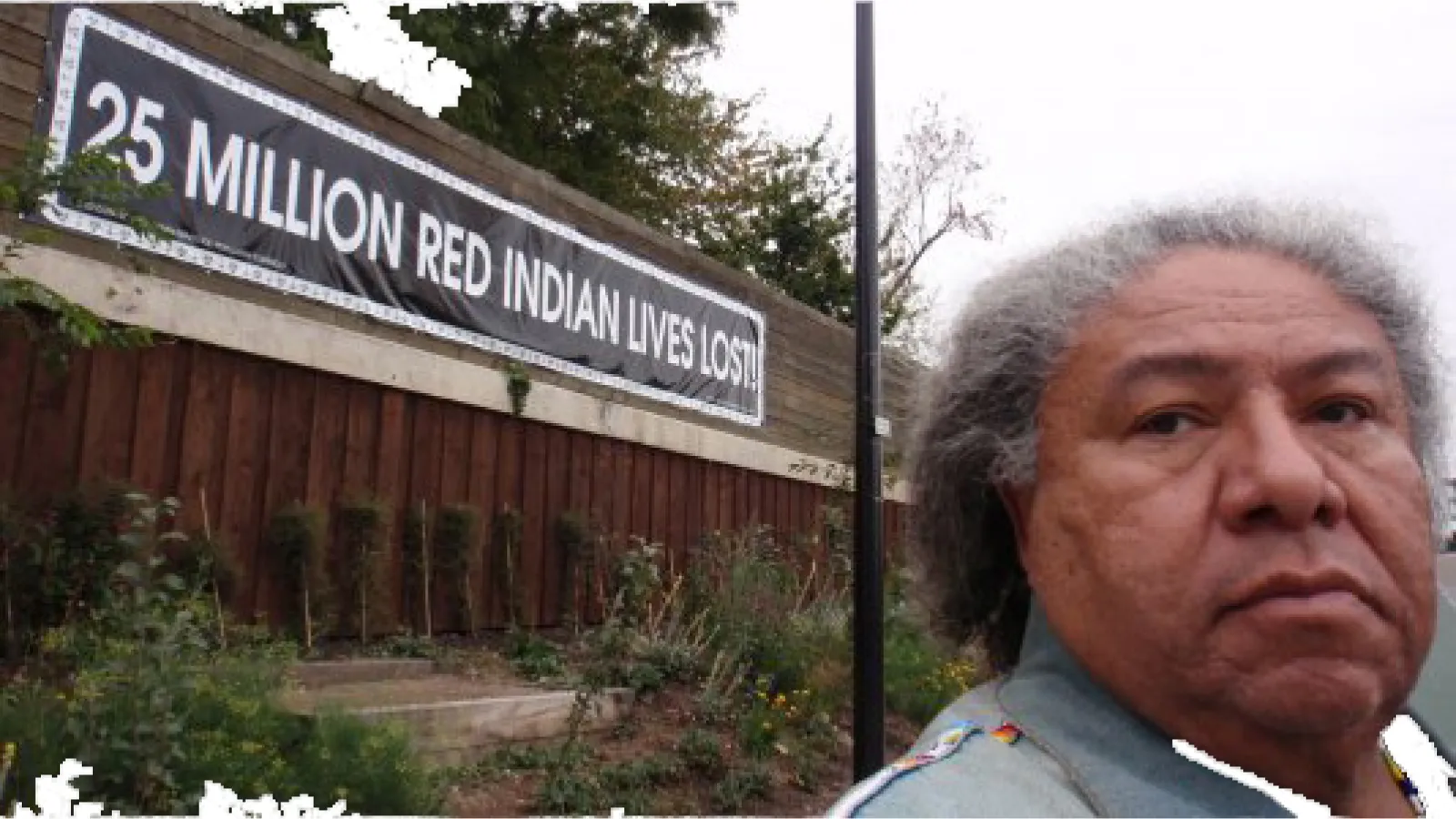 A brown skinned man with gray and white hair looks at the camera next to a sign on a building that says "25 million red indian lives lost"