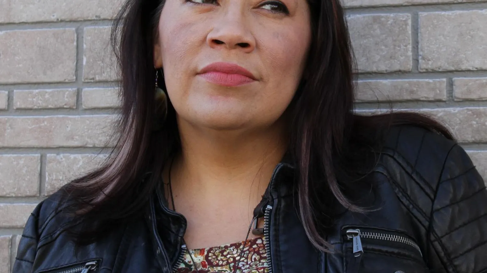 Kim Tallbear, a brown-skinned woman with long dark brown hair and wearing a multi-colored shirt and black jacket looks off to the left of the camera