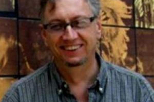 John Low in a blue shirt and glasses in front of a mosaic wall.