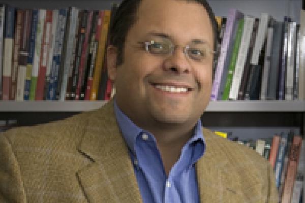 Bryan Brayboy, a tan-skinned man with glasses and a brown jacket sitting in front of a bookcase, smiling