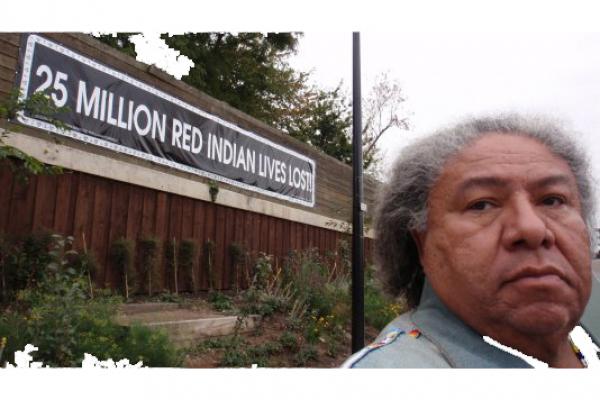 A brown skinned man with gray and white hair looks at the camera next to a sign on a building that says "25 million red indian lives lost"